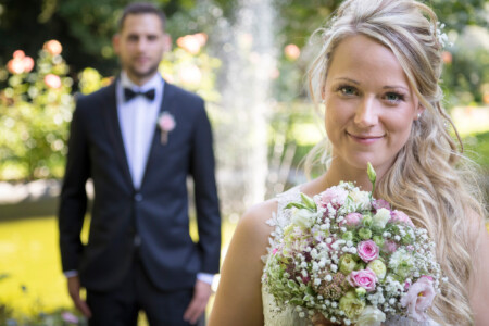 Braut scharf im Vordergrund, Bräutigam in der Unschärfe dahinter vor einem Springbrunnen Die Braut im Fokus